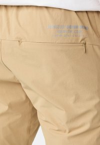 Pantalons légers beiges avec une texture subtile, dotés d'une poche arrière zippée et d'un texte de marque imprimé en bleu clair.