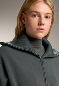 Tmavo zelená zipsová bunda s vysokým, rebrovaným golierom. Mäkká textúra a minimalistický dizajn. Šedý top nosený pod ňou, viditeľný pri výstrihu.