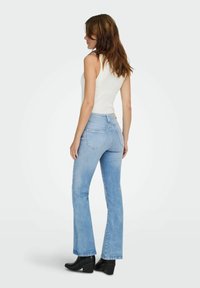 Šviesiai mėlynos plačios džinsinės kelnės su aukštu liemeniu, turinčios užpakalines kišenes ir lygią džinsinio audinio struktūrą, derinamos su balta marškinėlių tipo palaidine ir juodais aukštakulniais batais.