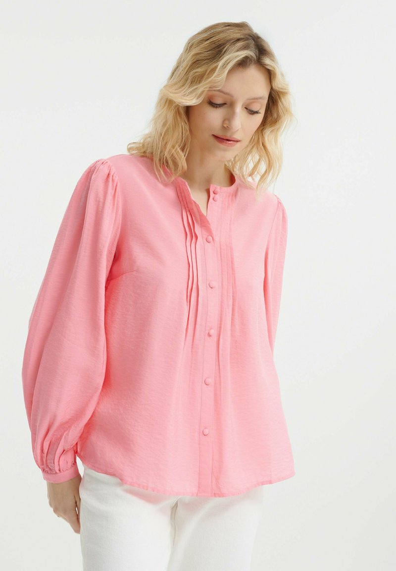 Hellrosa Bluse mit Knopfverschluss, langen Puffärmeln und vertikalen Falten an der Vorderseite. Hergestellt aus einem strukturierten, weichen Stoff.