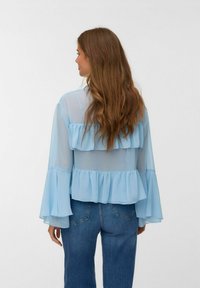 Ljusblå genomskinlig blus med lager av volangdetaljer, långa trumpetärmar och en lös passform, matchad med mörkblå denimjeans.