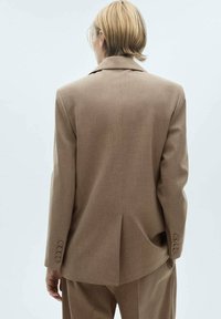 Personne aux cheveux courts et blonds, portant une veste de costume beige et un pantalon assorti, debout de dos sur un fond uni.