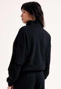 Sweatshirt noir à col montant et à manches longues. Doté d'un motif texturé le long des manches. Tissu doux, coupe décontractée.