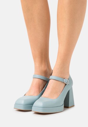 Femme portant des escarpins Mary Jane bleu clair à plateforme avec brides à la cheville et talons blocs, debout sur un fond blanc.