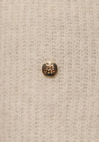 Cream-colored knit fabric with a soft texture features a round, metallic gold button embossed with "MOS MOSH" in a smooth finish.
