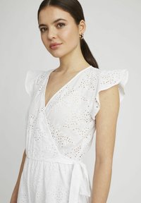 Robe en coton blanche avec broderie à œillets, décolleté en V, design portefeuille, manches flottantes et détail de taille à nouer, mettant en valeur un motif délicat.