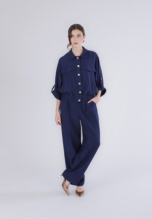 Mujer de cabello castaño viste chaqueta azul marino abotonada y pantalones anchos a juego, posando con una mano en el bolsillo y tacones sobre fondo blanco.