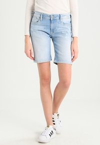 Pantalones cortos de mezclilla azul claro con un dobladillo deshilachado, diseño de cinco bolsillos y un ligero desteñido. Combinados con zapatillas blancas con rayas negras.