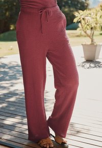 Pantalons larges maroon texturés avec taille réglable et coupe décontractée. Associés à des sandales noires, sur un fond de terrasse en bois.