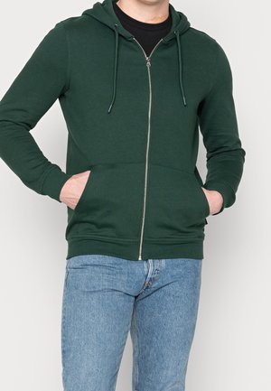Felpa con cappuccio verde con zip, realizzata in tessuto morbido, con cappuccio regolabile con coulisse, tasche frontali e polsini a coste. Abbinata a jeans azzurri.