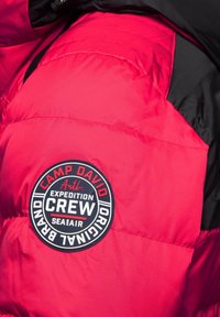 Chaqueta puff roja con acentos en negro, que presenta un parche de logo redondo que incluye "Camp David", "Expedition" y "Crew". Tejido texturizado.