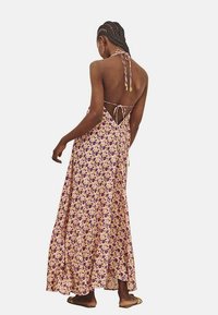 Vestido maxi hecho de una tela ligera, con un patrón floral en púrpura y amarillo sobre un fondo durazno, con cuello halter y espalda descubierta.