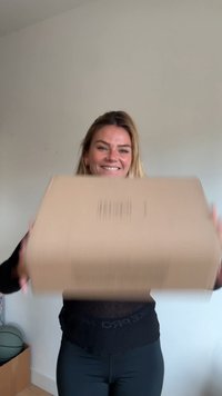 Caixa de cartão segurada com ambas as mãos; superfície castanha simples com vincos visíveis; sem rótulos ou marcas presentes. O fundo apresenta uma parede e alguns objetos.