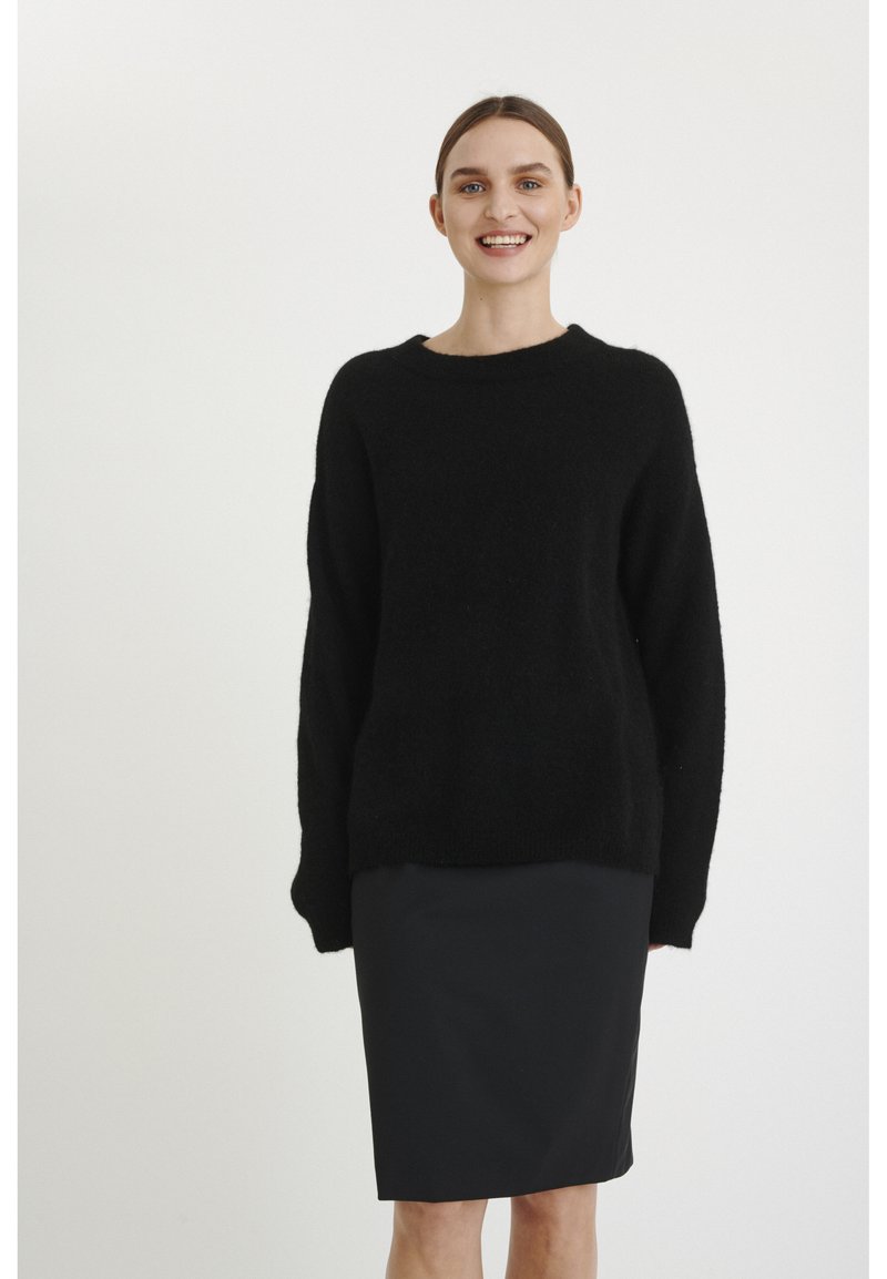 Maglione nero con scollatura tonda e maniche lunghe, realizzato in materiale morbido, abbinato a una gonna a tubino nera attillata, con una texture liscia.