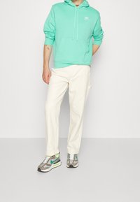 Mintgrön huvtröja med känguruficka, tillsammans med krämfärgade byxor och grå sneakers med gröna accenter och vit sula.