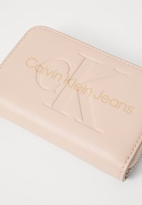 Calvin Klein Jeans piniginė iš minkštos rožinės dirbtinės odos, su iškilniu logotipu ir auksinės folijos užrašu, švariomis siūlėmis ir plonu dizainu.