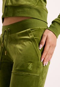 Zeleni velur športni komplet, ki vključuje krajši zgornji del in hlače s teksturirano obdelavo, vrvico za nastavitev pasu ter višje vezenje logotipa.