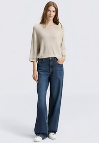 Top de punto beige con mangas tres cuartos combinado con jeans de pierna ancha azul oscuro de cintura alta y zapatillas blancas. Textura de tela lisa.