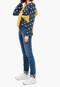Navy blouse with white polka dots, yellow knitted sweater tied around shoulders, blue jeans, and white sneakers, ankle cuffs rolled.