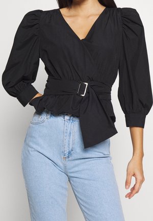 Frau trägt schwarze Wickelbluse mit Puffärmeln und Gürtel mit Schnalle, kombiniert mit hellblauen Jeans mit hoher Taille.