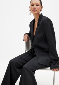 Črn krojen suknjič iz teksturirane tkanine, ki vključuje vezano zapiranje, široke hlače in rever z izrezom. Model sedi na kovinskem stolu.