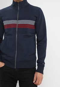 Giacca zip-up navy con colletto alto, presenta righe orizzontali in rosso, bianco e blu sul petto, realizzata in un tessuto testurizzato.