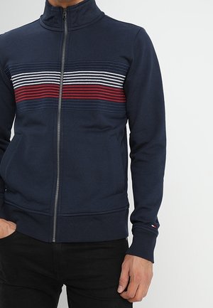 Giacca zip-up navy con colletto alto, presenta righe orizzontali in rosso, bianco e blu sul petto, realizzata in un tessuto testurizzato.