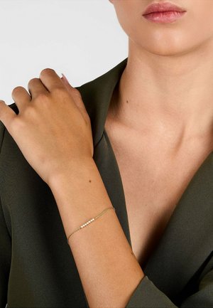 Woman's hand resting on shoulder, wearing a delicate gold bracelet with small pearls, dressed in a dark green V-neck garment.