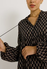 Vestido negro con estampado y corte holgado, que cuenta con un cuello, cintura fruncida y mangas largas abullonadas, adornado con motivos en dorado claro.