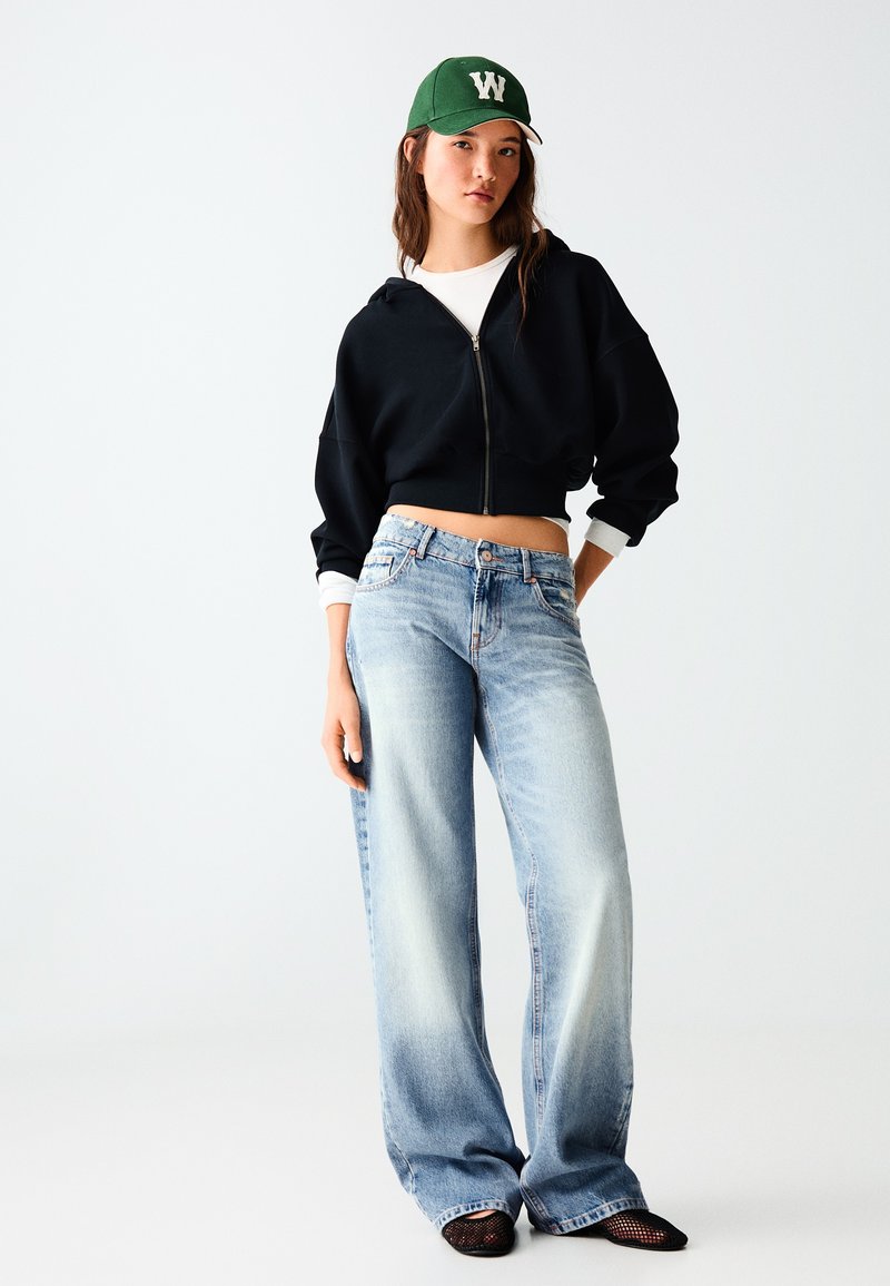 Jeune femme portant une casquette verte, un sweat à capuche court noir, une chemise blanche, un jean large bleu clair et des chaussures en maille noires, debout devant un fond blanc.