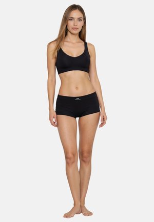 Donna in piedi a piedi nudi che indossa un reggiseno sportivo nero e pantaloncini attillati neri su uno sfondo bianco semplice.