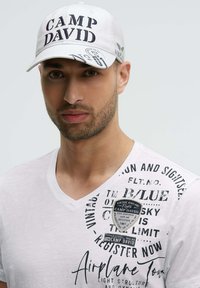 White cap with "CAMP DAVID" embroidered in black, features logos and text. Model wears a white T-shirt with printed text and patches.