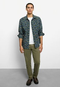 Marineblaues Hemd mit Blumenmuster und Knopfleiste, getragen über einem weißen T-Shirt. Kombiniert mit olivgrünen Hosen und dunkelbraunen Schuhen.