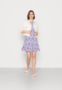 Vestido azul-claro com padrão floral vermelho e bainha com babados, combinado com uma jaqueta de ganga branca cropped e sandálias brancas. Fundo minimalista.