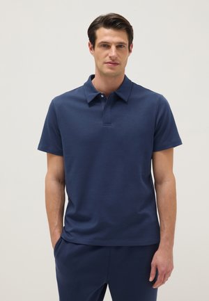 Hombre vestido con camisa polo de manga corta azul marino y pantalones a juego, de pie con las manos en los bolsillos contra un fondo claro y liso.