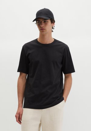 Młody mężczyzna w czarnej czapce i czarnym krótkim rękawie koszulce, stoi z jedną ręką w kieszeni kremowych spodni na jednolitym tle.