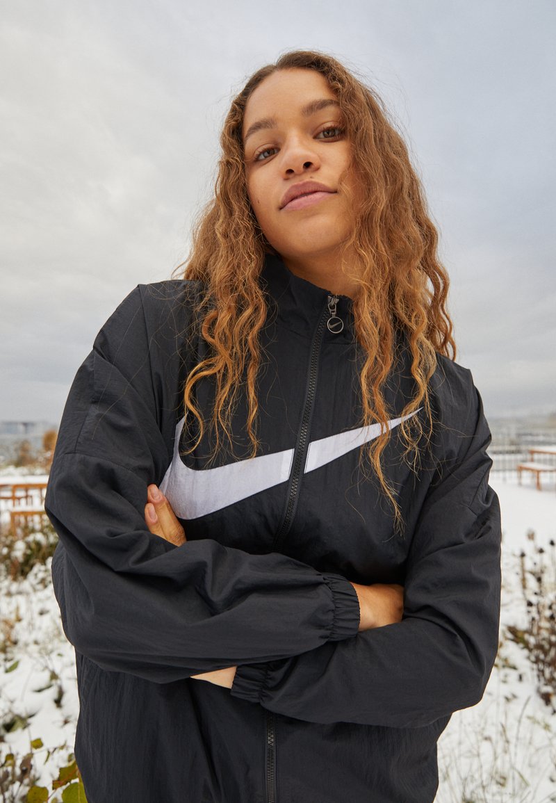 Musta tuulitakki, jossa on näkyvä valkoinen Nike-swoosh-logo. Varustettu ylösnostettavalla kauluksella ja elastisilla hihoilla, kevyttä materiaalia.