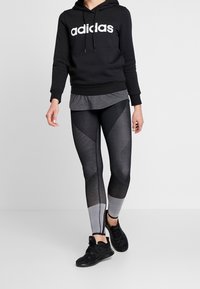 Persona che indossa una felpa nera Adidas, leggings sportivi neri e grigi con un motivo e scarpe da ginnastica nere Adidas mentre cammina su uno sfondo chiaro e uniforme.