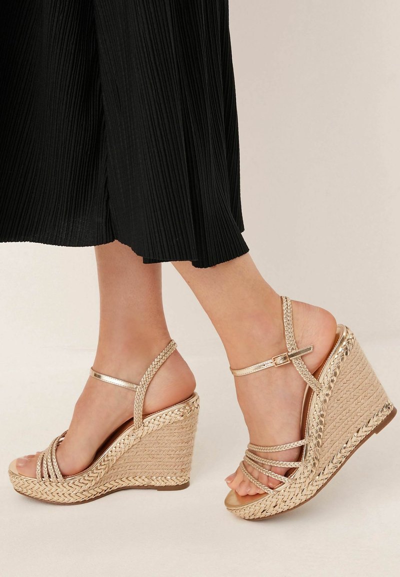 Pieds portant des sandales compensées dorées à brides avec des détails tressés, associées à un pantalon noir plissé court sur un fond clair.