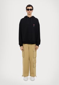 Felpa nera con cappuccio e tasca frontale, impreziosita da ricamo di un lecca-lecca rosso, abbinata a pantaloni cargo beige con più tasche e sneakers bianche.