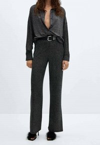 Femme portant un blouse pailleté gris anthracite avec un décolleté en V profond, un pantalon large assorti, une ceinture noire et des sandales à talons hauts avec bordure en fourrure noire.