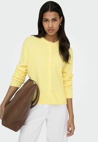 Rumeno jopico z gumbi, z rebrastimi manšetami in spodnjim robom. Kombinirana z rjavo usnjeno torbico, postavljeno na nevtralno ozadje.