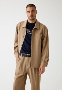 Camicia button-up color sabbia con colletto, indossata sopra una maglietta blu navy con testo bianco. Pantaloni beige e una cintura nera completano l'outfit.