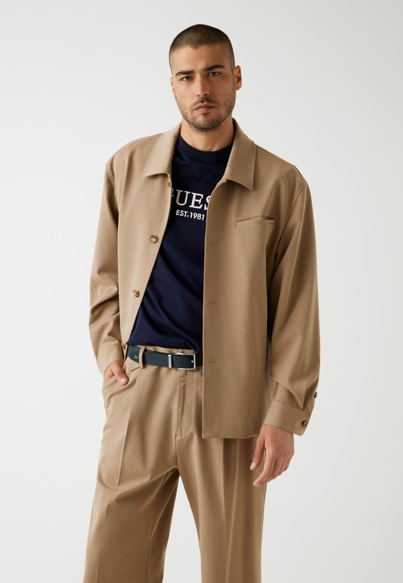 Camicia button-up color sabbia con colletto, indossata sopra una maglietta blu navy con testo bianco. Pantaloni beige e una cintura nera completano l'outfit.