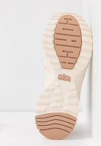 Semelle de chaussure beige et marron clair avec motif de grip texturé et petit logo tracteur au centre, inclinée sur fond blanc.