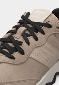 Μπεζ σουέντ αθλητικό παπούτσι με μαύρα κορδόνια, ορατές ραφές, με το εμπορικό σήμα "Sorel" στην πλευρά και στη γλώσσα, και μέρος της λευκής σόλας με μαύρη λεπτομέρεια.