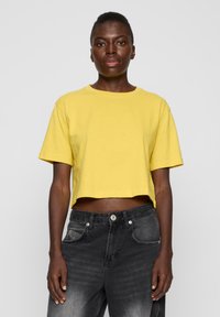 Kuttet gul bomulls T-skjorte med korte ermer, rund hals og en avslappet passform, sammen med svarte denimjeans med falming.