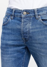 Ljusblå denimjeans med en figursydd design, synliga sömmar och en metallknapp i midjan. Två framfickor ingår.