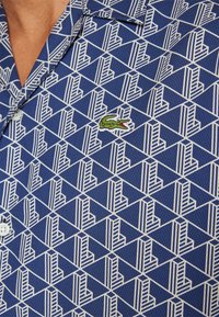 Camisa de manga corta con un patrón geométrico en azul y blanco, que presenta un logo de cocodrilo bordado en verde en el pecho.