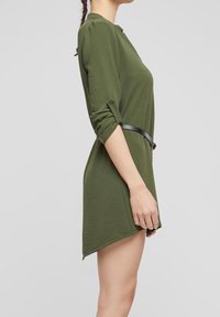 Femme en robe asymétrique verte à manches longues, manches retroussées et fine ceinture noire, vue de profil sur un fond uni.